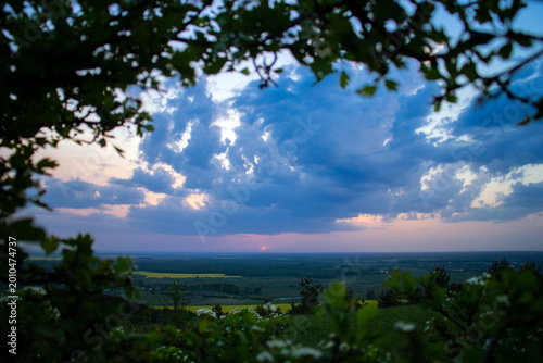 Photography Dramatic cloudy sky at sunset over a vast green countryside landscape framed by tree branches