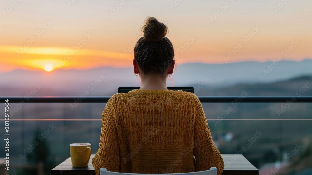 Fototapeta premium A tranquil scene of a woman working remotely on her laptop from a balcony during a vibrant sunset with a scenic mountain view and a warm drink embodying a peaceful lifestyle