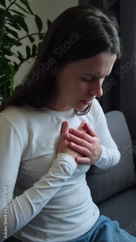 Close up of suffering acute cardiac young caucasian woman hand touching on her chest pain, female breathing problem, pressure sitting alone on couch at home. Heart attack, health care concept