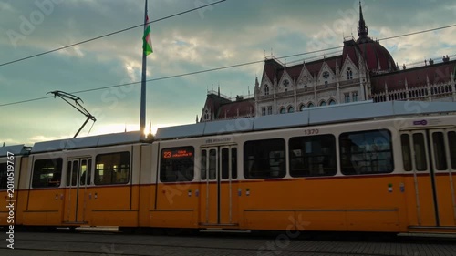 Yellow tram moving past the Hungarian Parliament Building in Budapest during a golden sunset with a tall flagpole and cloudy sky