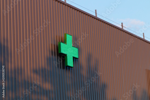 Illuminated green pharmacy cross sign on a corrugated brown building exterior