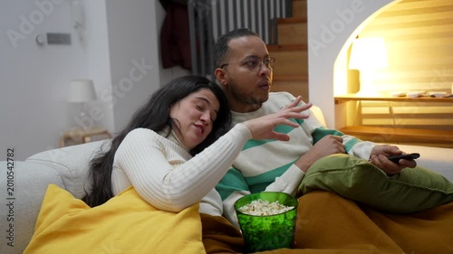 Happy couple watching a movie together on the sofa at night, eating popcorn and enjoying their leisure time at home. The man uses the remote control while the woman cuddles up to him