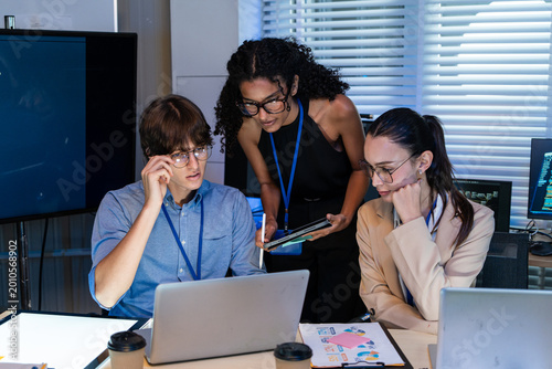 Serious multiracial tech team focused on laptop screen while working in office. Young professional colleagues using digital tablet and computers for software development during group project meeting