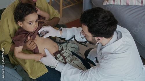 View from above of male multiethnic paediatrician in white coat using stethoscope while examining small patients breath sitting with mom during home appointment