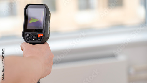 Hand grips a black thermal camera. Screen shows color-coded temperature patterns. Blurred building with windows in the background. Professionals use this for energy audits
