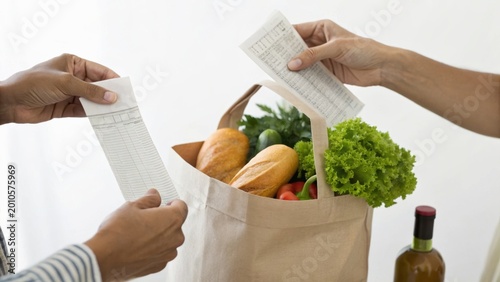 Two people holding grocery receipts near a paper bag filled with fresh vegetables and bread, with a bottle of olive oil nearby on a white surface.