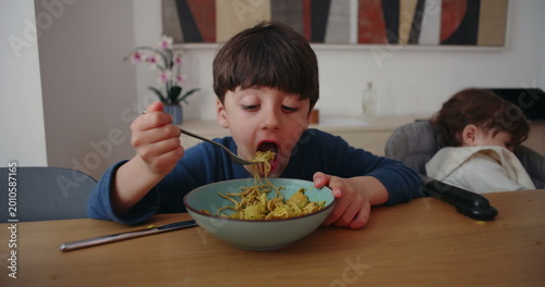 Fotografija Young boy eating noodles with fork at dining table while baby sibling sleeps in