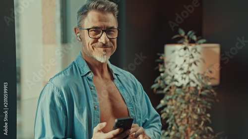 Mid adult man standing in sunlight in open shirt in the morning, checking email, news or social media on cell phone. Happy older male smiling in glasses.