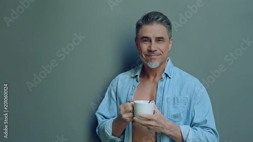 Confident smiling mature man holding a coffee mug, standing in front of a gray wall, looking at camera. Mid adult male in 50s, open denim shirt, bearded, grey hair, friendly and approachable smile.