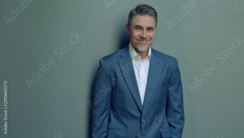 Well-dressed man in 50s standing against neutral backdrop, confidence and approachability with warm, charming smile. Business portrait of happy, friendly mature businessman smiling.