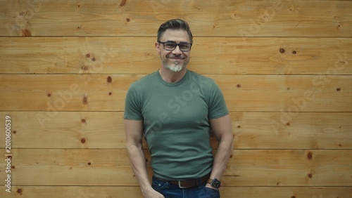 Portrait of happy mature man with eyeglasses and casual clothes standing, leaning against wooden wall, smiling. Mid adul, middle aged male in 50s. Copy space.