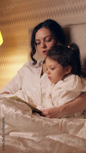 Caring mother reading a book to her sleepy little daughter in bed. Loving family enjoying a quiet evening together with a fairytale