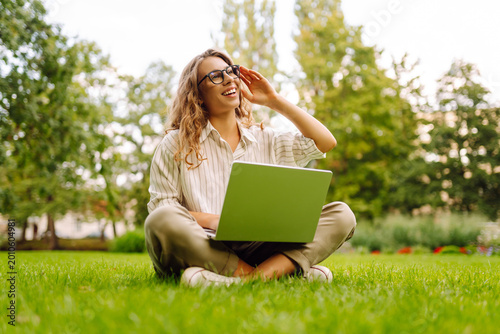 Fotografie A happy woman sits on the grass in a park with a laptop on a sunny day