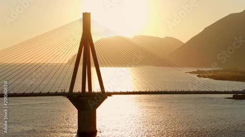 Famous Rio-Antirrio bridge silhouetted against a golden sunset, with shimmering water reflections and distant mountains creating a breathtaking and scenic hellenic landscape
