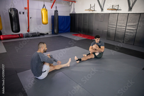 Diverse male training partners stretching on mats in gym wearing athletic shorts with ankle tape