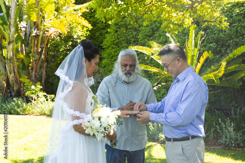 Couple with Indian officiant exchanging rings on sunlit lawn, bride holding white bouquet and veil