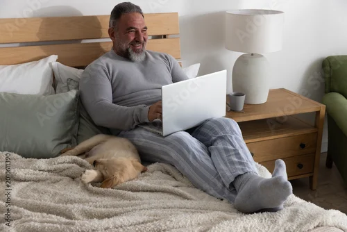 Fototapeta Senior man sitting on bed typing on laptop in pajama pants with sleeping dog and mug