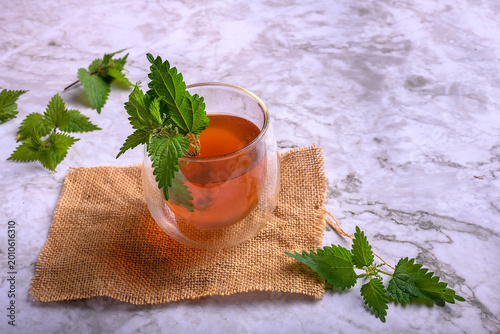 Fotografi Nettle herbal tea in a double wall glass on burlap and marble surface, fresh sti