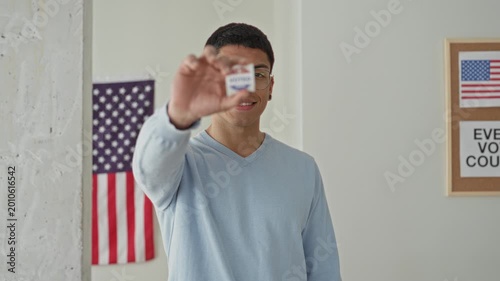 Young hispanic man holds 'voted' sticker between fingers, smiling, in a polling building with american flag visible; civic pride.