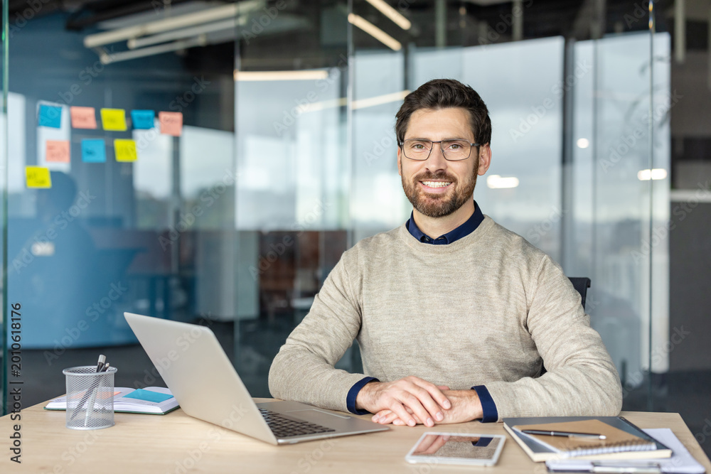 Fototapeta premium Smiling bearded professional wearing glasses sits at a modern desk with laptop and office supplies, looking confident and relaxed while working remotely in a contemporary workspace