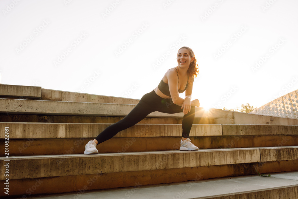 Fototapeta premium An athletic woman exercises on the steps on a sunny day. A young woman in athletic clothing stretches while exercising outdoors at sunset. Concept of activity, sports, and weekends.