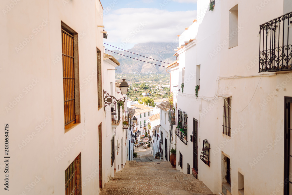 Obraz premium A narrow street leads through a small village with white buildings on both sides. The area has plants on balconies and mountains in the background under a clear sky.