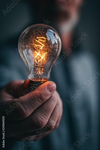 A close up view of a man's hand holding a glowing incandescent light bulb, symbolizing ideas and innovation.