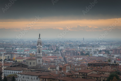 Bergamo skyline with statue-topped clock tower and airplane beneath dramatic cloudy sky
