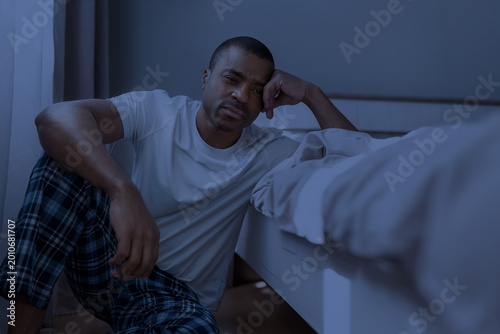 Sleepless African American man in pajamas sitting on bedroom floor beside bed at night, leaning with troubled expression in dark blue light. Insomnia and stress concept