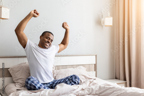 Cuadro en lienzo African american man stretching arms while sitting in bed in bedroom