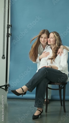 Vertical video of beautiful mature mother and adult daughter in white shirts and jeans hugging in the photostudio on blue paper background. Two female generations. Mother's day concept