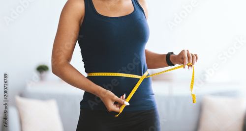 A woman stands indoors and uses a yellow measuring tape to measure her waist. She focuses on her body measurements as part of her fitness routine. The setting is bright and open.