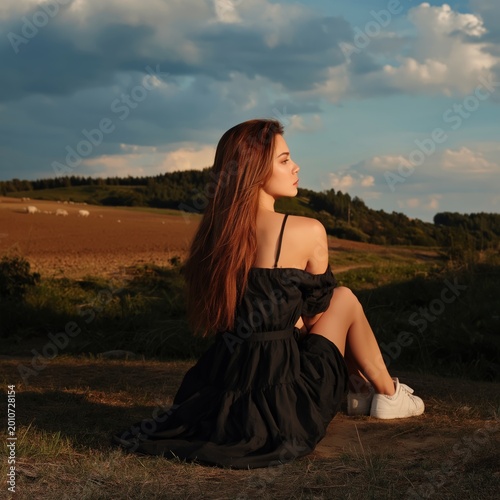 Woman Sitting on Grass in a Field During Sunset Near a Forest and Clouds