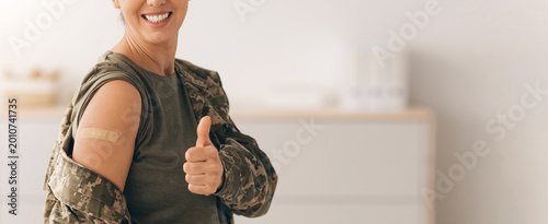 A woman in military attire smiles and gives a thumbs up after getting a vaccination. She has a band-aid on her arm and appears happy in a medical facility.