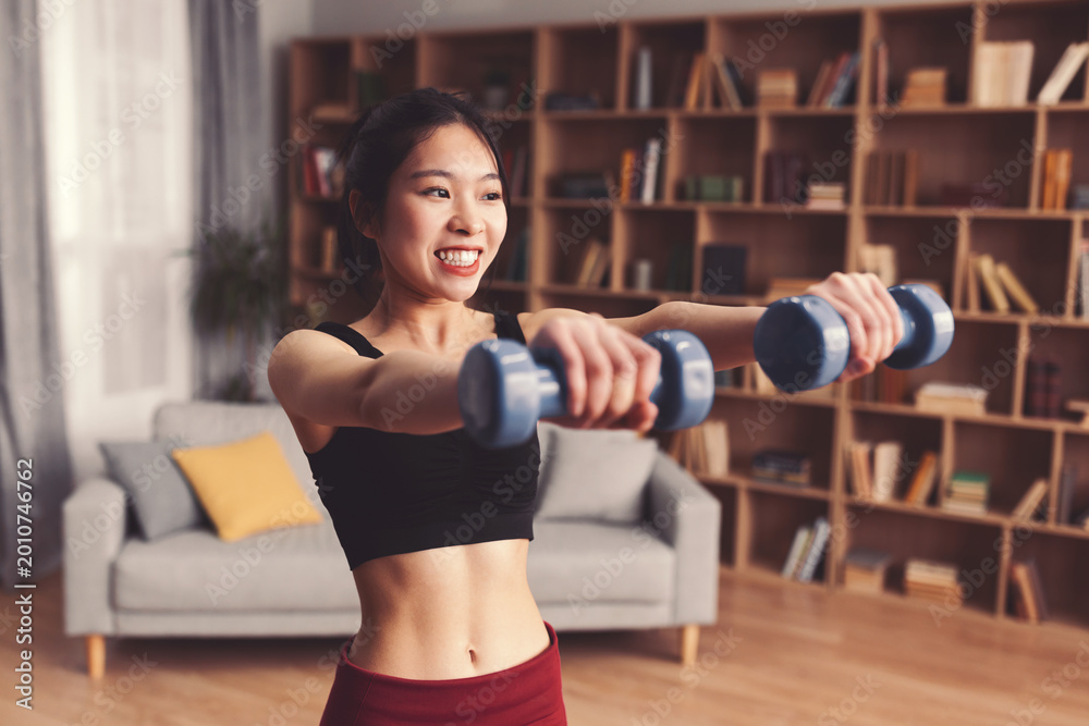 Naklejka premium A woman is exercising in her living room. She holds dumbbells while smiling and focusing on her workout. The space has a couch and a bookshelf. Morning light shines through the window.