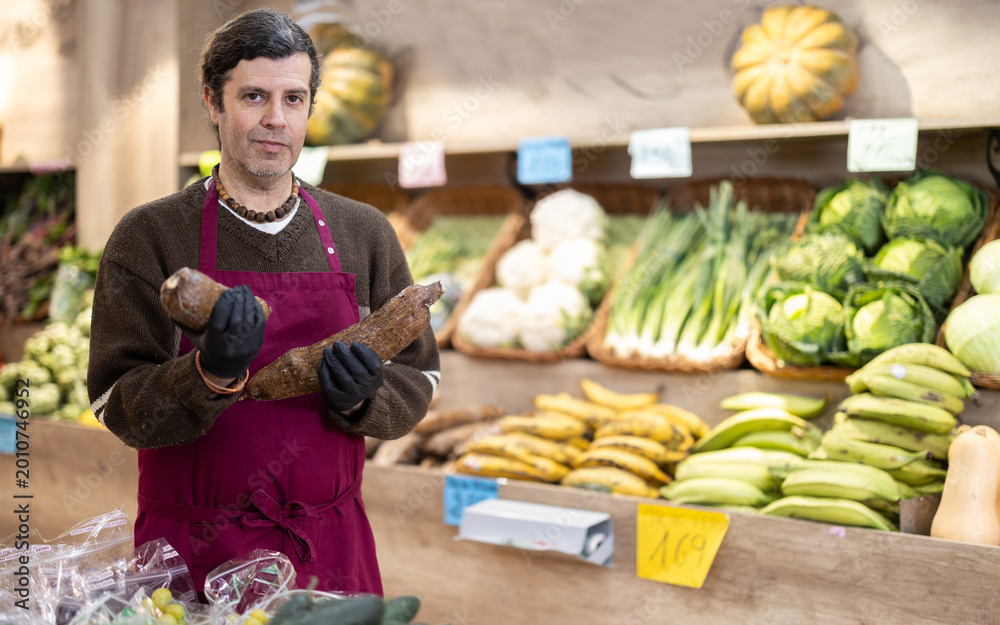 Fototapeta premium Adult man seller in apron with cassava in vegetable shop