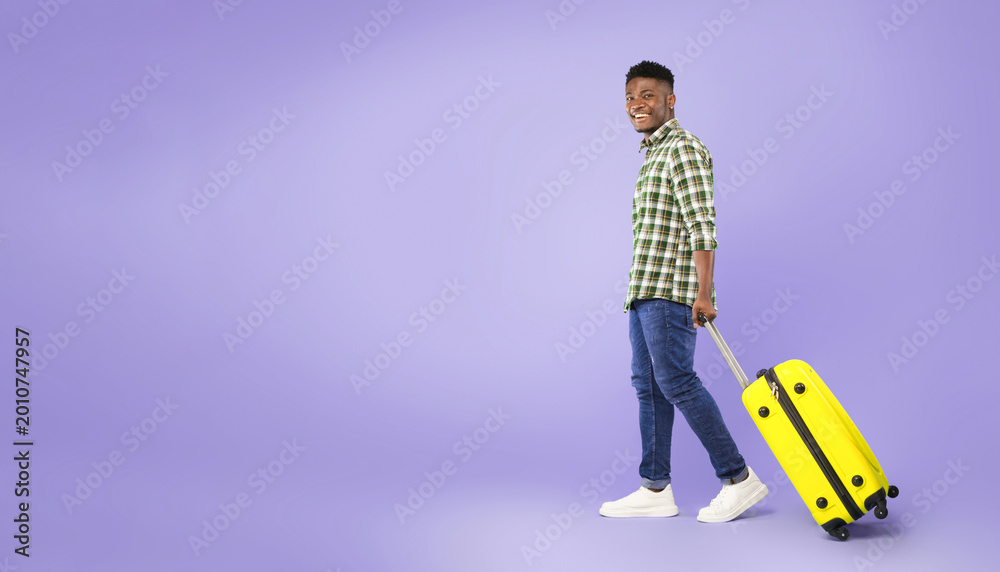 Fototapeta premium A young man is walking while pulling a bright yellow suitcase. He is smiling and dressed in checkered clothing. The background is a solid purple color, suggesting a fun and vibrant atmosphere.