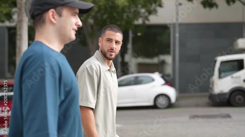 Two men walk side by side on a paved street. They wear casual attire, one with a cap. Their expressions are neutral, focused on forward motion. Delivery truck rests parked nearby