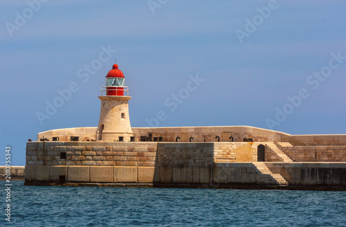 Stone lighthouse with red lantern on the breakwater in Grand Harbour of Valletta, Malta. Historic maritime structure on the Mediterranean coast