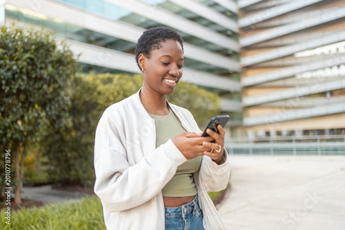 Smiling young businesswoman in smart casuals using smartphone while standing near modern office building. Focused expression and digital communication concept