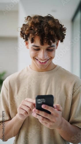Young man in beige t-shirt smiling while using a smartphone indoors medium shot