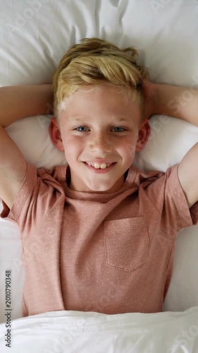 Blonde boy in pink shirt lying on white bed sheets smiling at camera high angle view