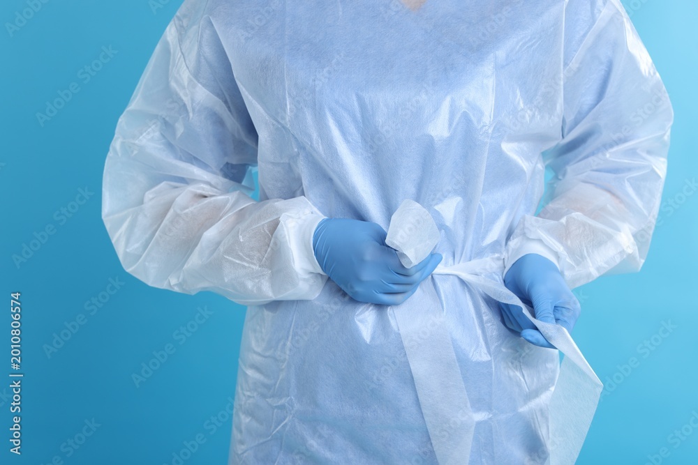 Fototapeta premium Medical worker tying disposable isolation gown on light blue background, closeup