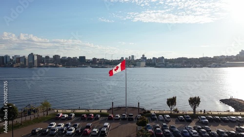 Cinematic drone view of Canadian flag waving on Dartmouth waterfront with Halifax skyline glowing in warm summer sunset over Atlantic Ocean. Bridge and downtown visible, symbol of national pride 
