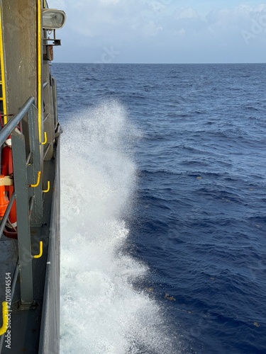 Close side perspective of a moving ship showing intense sea spray hitting along the hull, deep blue ocean water, and visible structural elements of the vessel, highlighting speed, force