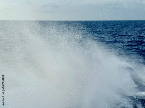 Abstract view of ship wake trailing behind a moving vessel, showing foamy white water patterns blending into deep blue ocean, with soft horizon line creating minimalistic maritime background