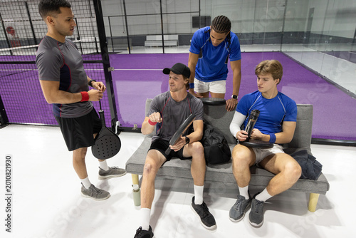 Photography Male teammates in sportswear sitting on bench at purple court, holding paddles w