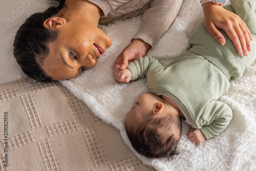 African American mother lying on plush blanket at home, touching infant hand, smartwatch visible