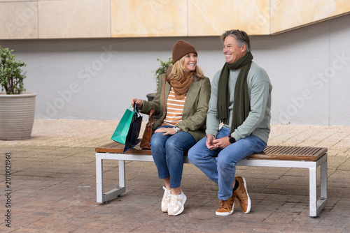 Senior couple sitting on bench at paved plaza holding shopping bags and handbag, wearing scarves