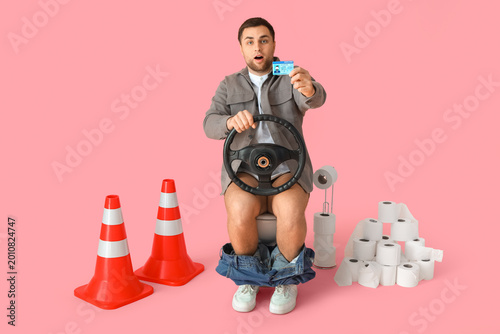 Shocked driver with license and steering wheel sitting on toilet bowl against...